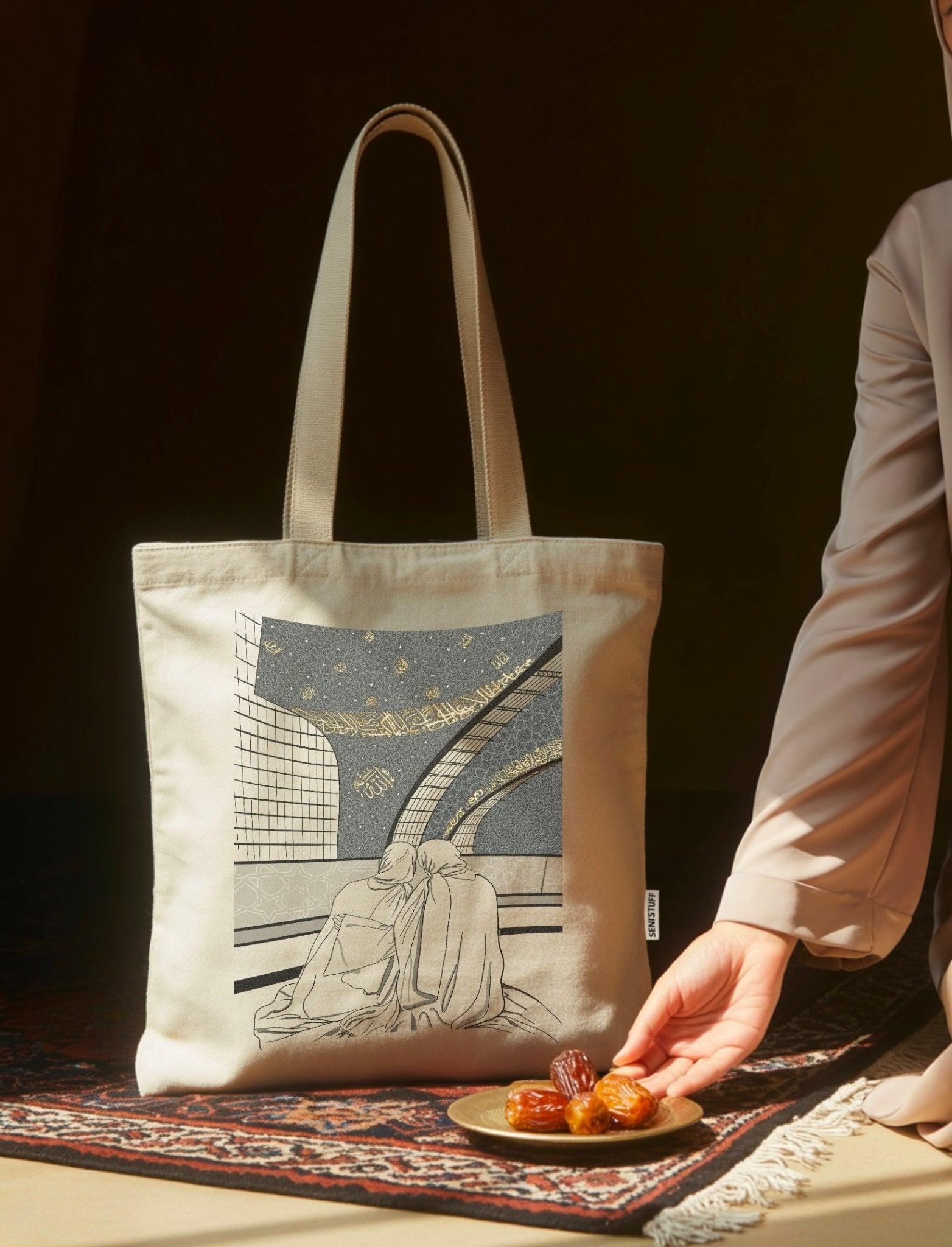 Sac en toile coton beige avec dessin de deux femmes en prière dans une mosquée, présenté avec une assiette de dattes sur un tapis oriental.