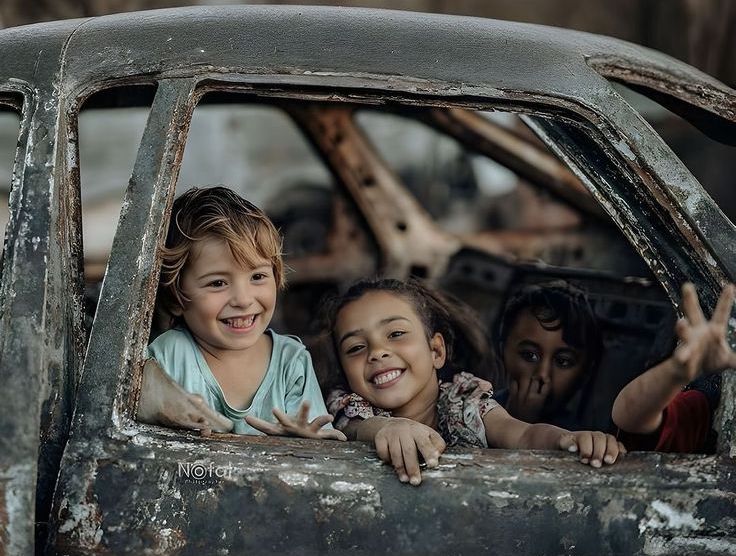 Enfants palestiniens souriants assis dans une carcasse de voiture détruite, symbole d’espoir et de résilience malgré la guerre.