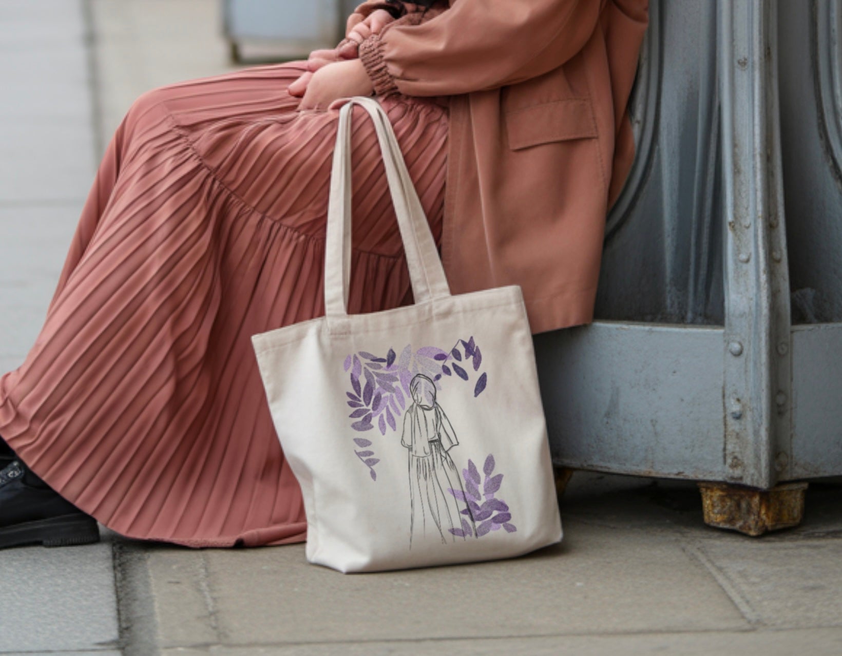 Tote bag coton avec illustration minimaliste de femme voilée entourée de motifs floraux violets, posé au sol à côté d’une jupe plissée rose.