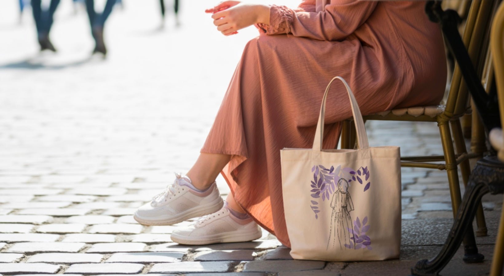 Tote bag coton beige avec dessin de femme et feuillage violet, posé au pied d’une table de café en extérieur, ambiance lifestyle chic.