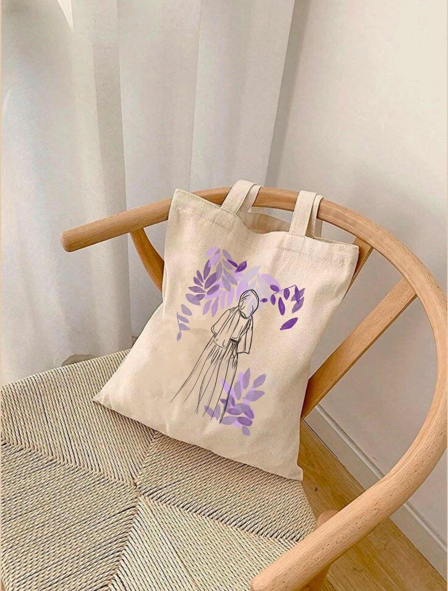 sac en toile Seni'Stuff avec dessin minimaliste de femme voilée et feuilles violettes, posé sur une chaise près d’un rideau