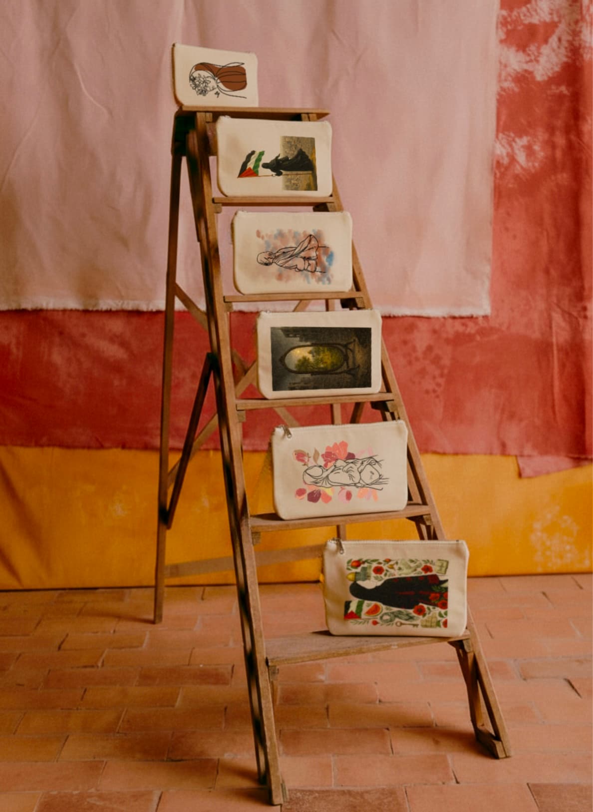 Six pochettes illustrées Seni'Stuff disposées sur une échelle en bois devant un fond textile rose et ocre, évoquant une galerie artisanale et poétique.