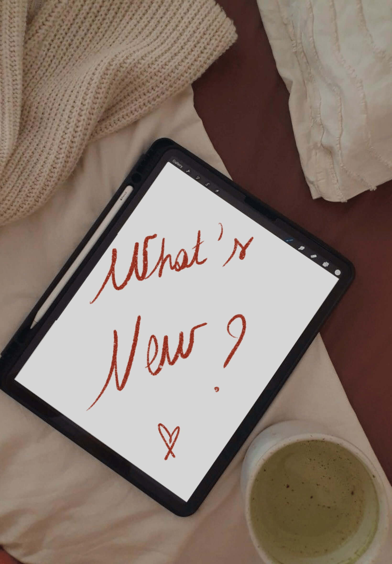 Une tablette affichant le message manuscrit “What’s New?” posée sur un lit beige avec un matcha, ambiance cocooning et créative