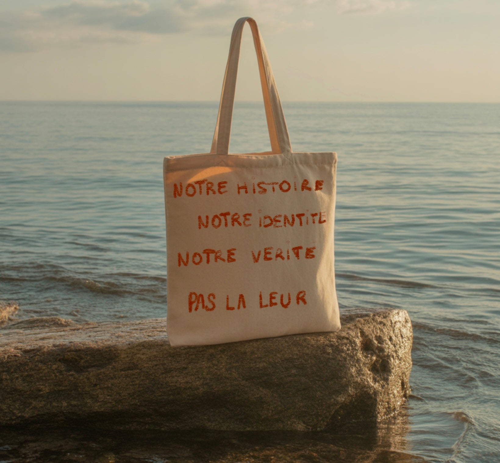Sac cabas en toile beige avec inscription rouge « Notre histoire, notre identité, notre vérité, pas la leur », posé sur un rocher au bord de la mer.