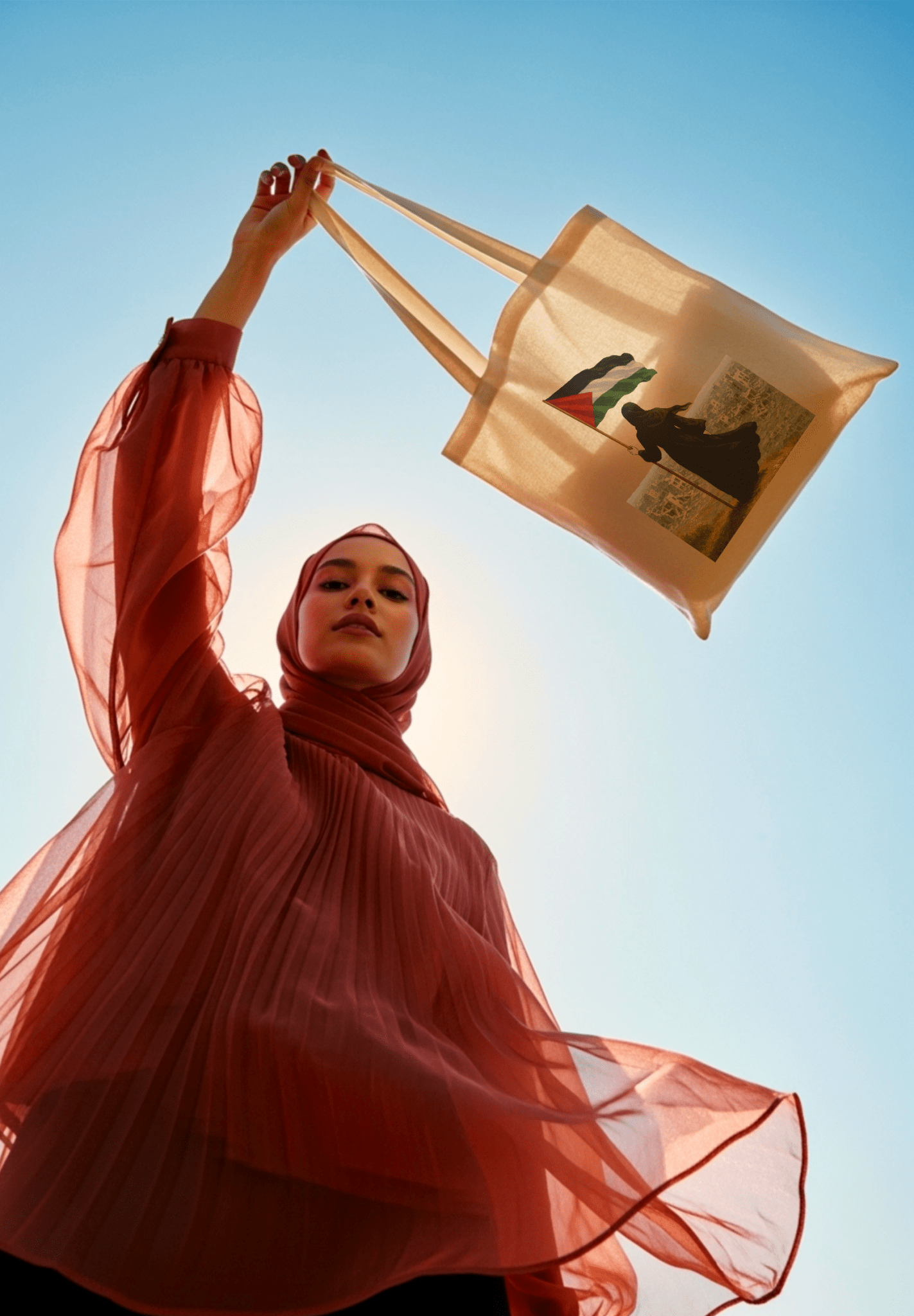 Femme voilée en hijab terracota levant un sac en toile beige vers le ciel bleu avec l'illustration d'une femme voilée qui tient le drapeau de la palestine dans la main face aux décombres de la ville 