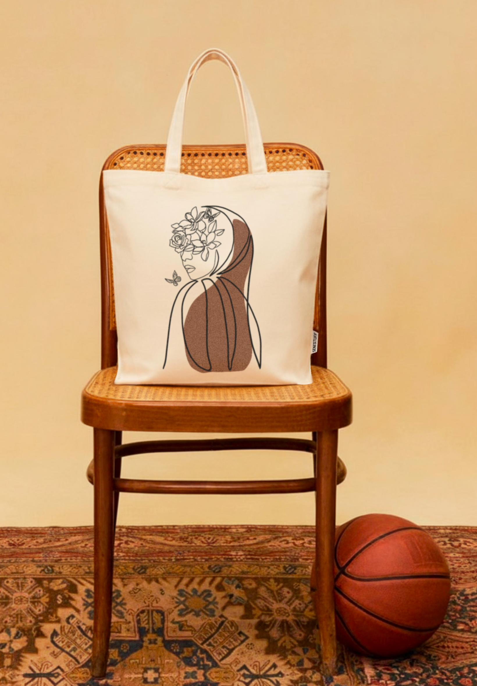 Sac en toile beige Seni’Stuff avec illustration d’une femme voilée entourée de fleurs, posé sur une chaise vintage en bois à côté d’un ballon de basket.